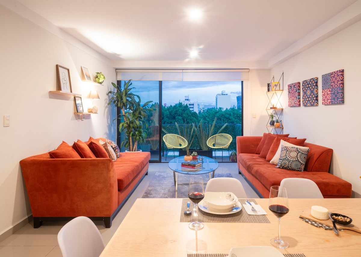 A modern living area features two vibrant orange sofas positioned around a glass coffee table. The space is adorned with green plants and framed artworks on the walls. Large windows invite natural light and offer views of greenery outside, enhancing the atmosphere.