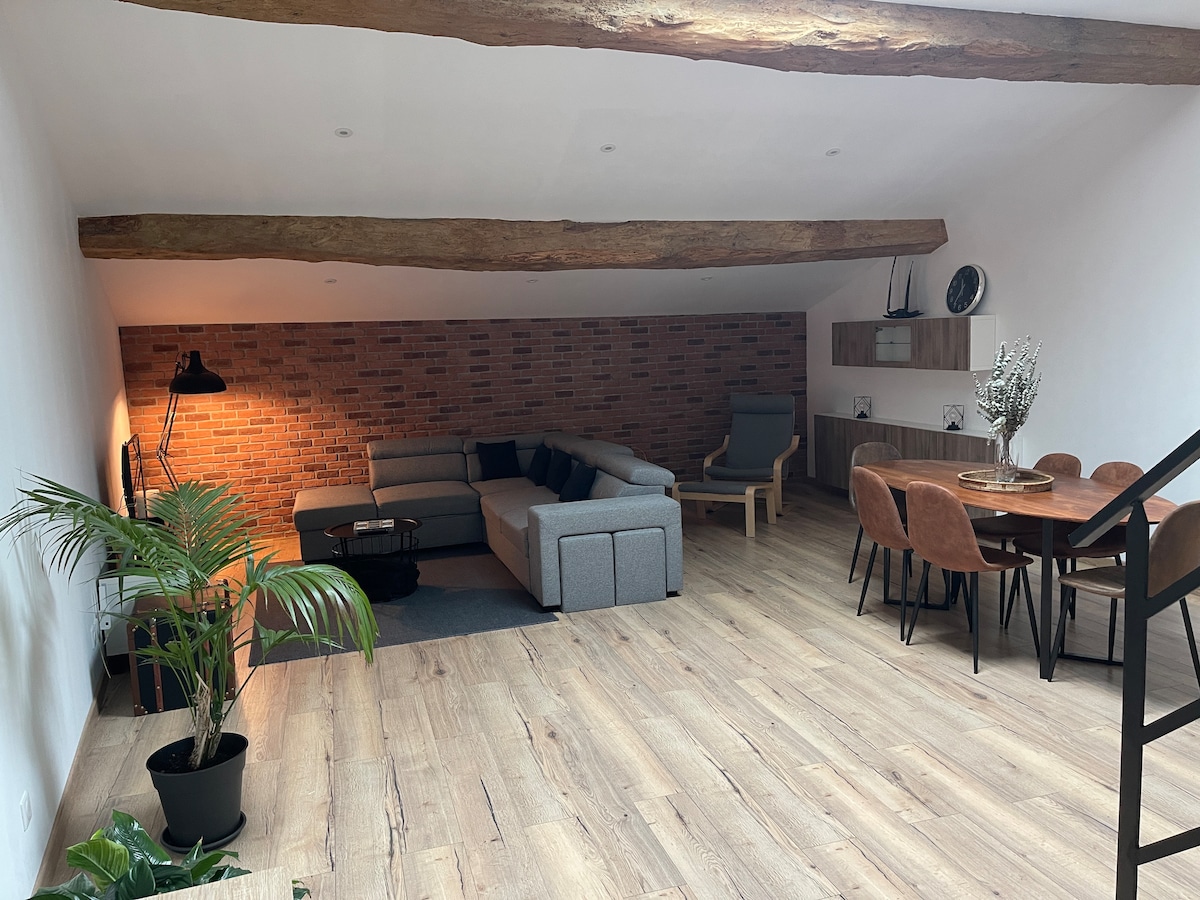 A spacious living area features a sectional sofa arranged around a low table, with a wooden beam ceiling adding character. A dining table with chairs is visible in the background, complemented by a soft light from a floor lamp. A potted plant adds a natural touch.