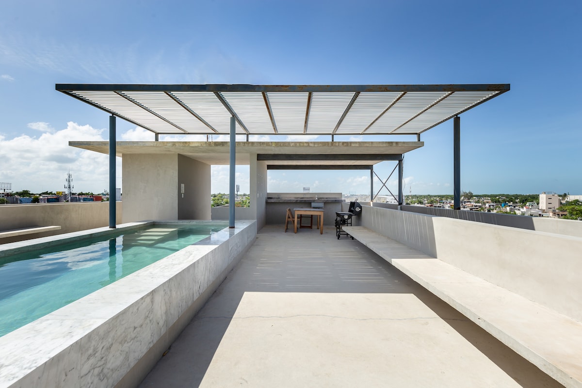 A rooftop terrace offers a spacious area with a swimming pool and marble seating. A wooden table is set up for outdoor dining. The surrounding skyline and views are visible under a clear sky.