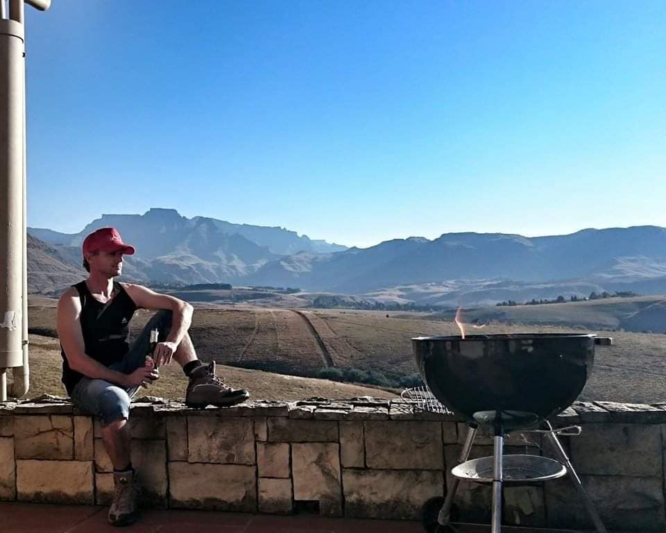 A person sits on a stone wall overlooking vast rolling hills and a mountain range in the distance. A barbecue grill is positioned nearby, with gentle flames visible. The scene is bathed in bright sunlight, creating a warm ambiance.
