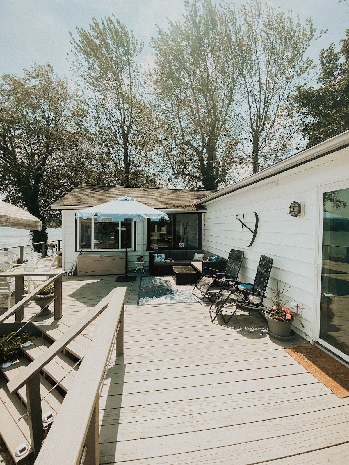 A spacious deck is visible, featuring comfortable seating options under a blue umbrella. A sliding glass door leads into the home, surrounded by greenery and trees. Potted plants add a touch of color, with a serene view of the water in the background.