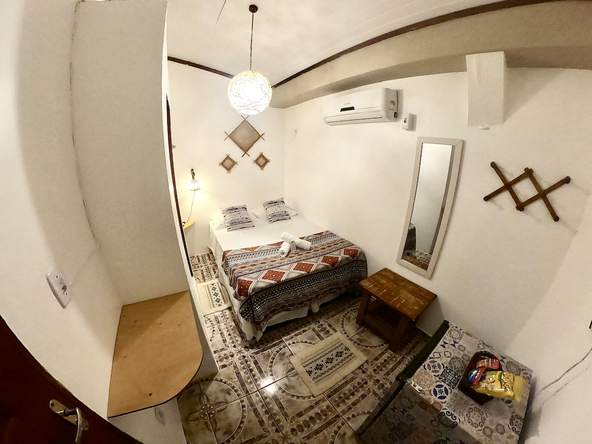 The cozy suite features a spacious bed covered with a patterned blanket. A small wooden table and a mirror are positioned nearby. Decorative elements adorn the walls, and natural light filters through the room, creating an inviting ambiance.