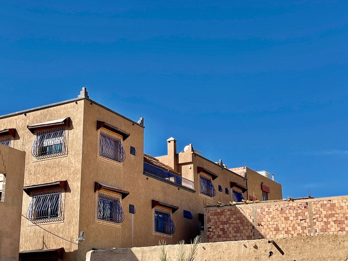 The exterior of the guest house is displayed against a clear blue sky. The building features warm earth-toned walls and distinctive decorative window grilles. A stone wall is visible in the foreground, which enhances the overall architectural character of the property.
