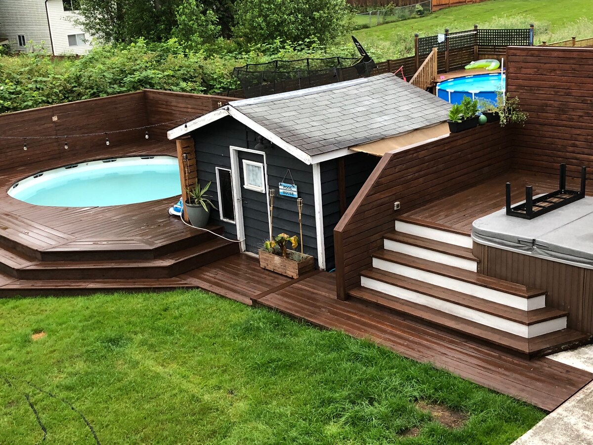A private outdoor area features a circular pool surrounded by wooden decking. A small shed with a gray exterior sits adjacent to the pool. Steps lead to a raised platform, adding depth to the landscape. Lush greenery and a wooden fence provide a natural backdrop.