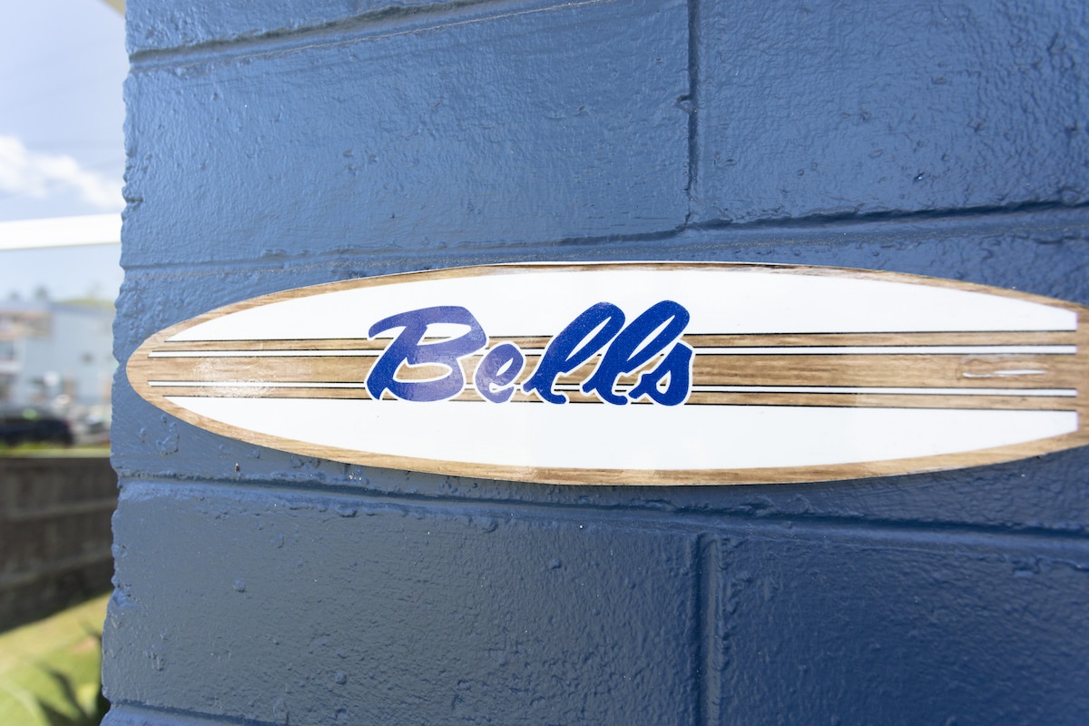 A wooden sign with the word 'Bells' painted in blue is affixed to a textured blue wall. The sign is shaped like a surfboard, emphasizing the coastal theme of the location.