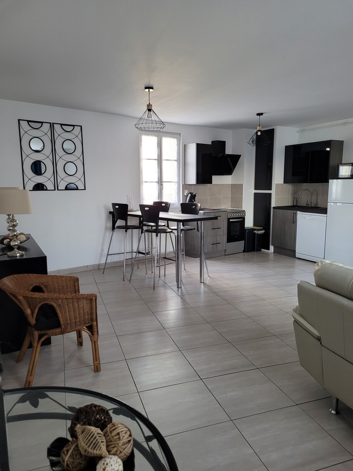 An open-plan kitchen and dining area is featured, showcasing a modern design with a tall table and high stools. Kitchen appliances include a refrigerator and stove. A woven chair adds comfort, while natural light enters through a window, illuminating the tiled floor.