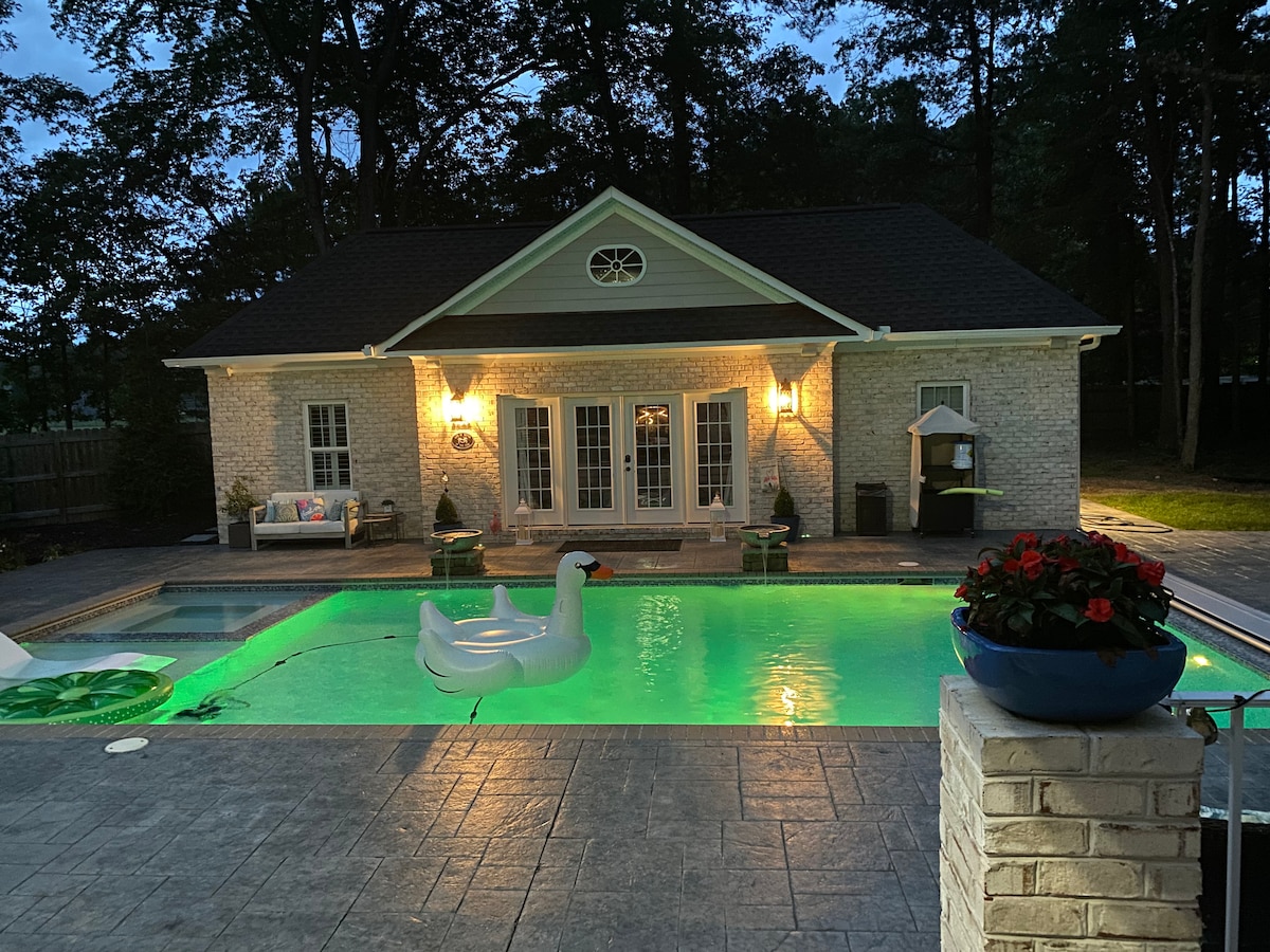 The exterior features a charming house illuminated by soft lighting, positioned beside a tranquil pool. A large white float shaped like a swan is seen in the green-tinted water. Lush landscaping surrounds the area, creating a serene outdoor retreat.