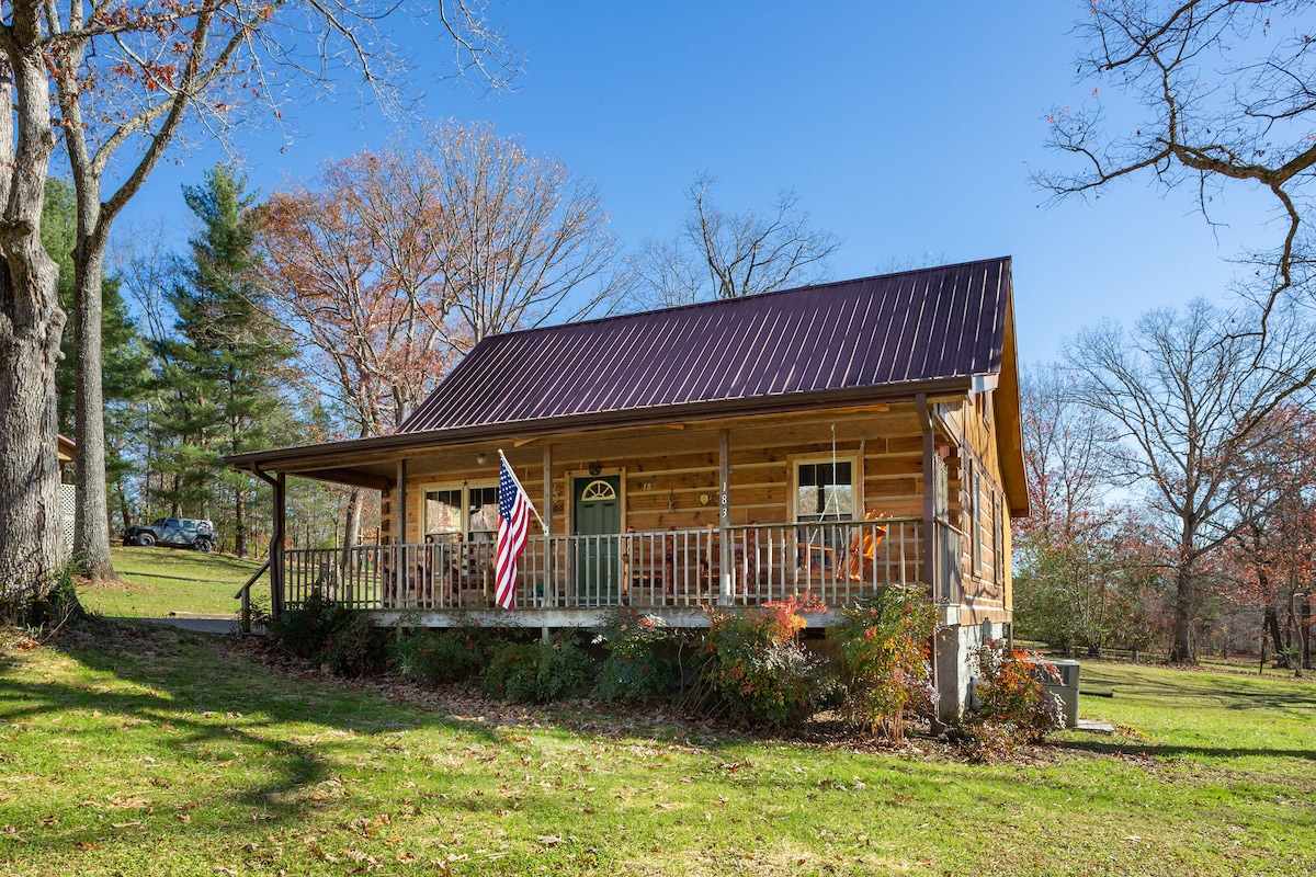 The cabin features a welcoming front porch with railings, flanked by rocking chairs and a swing. An American flag is prominently displayed. The cabin’s wooden facade is complemented by a metal roof, and lush greenery surrounds the property, creating a serene outdoor space.