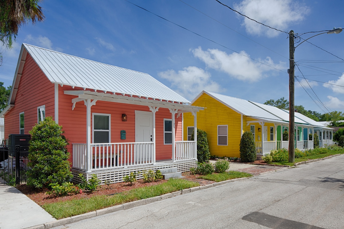 Colorful cottage-style homes are displayed along a tree-lined street, with a vibrant pink cottage on the left and a cheerful yellow cottage on the right. Each features a welcoming porch and a well-maintained front yard, under a clear blue sky.