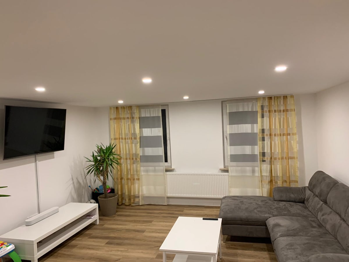 A modern living area is presented, featuring a gray sectional sofa beside a bright white coffee table. An entertainment unit holds a large flat-screen television. Natural light enters through two windows adorned with light curtains, while a potted plant adds a touch of greenery to the space.