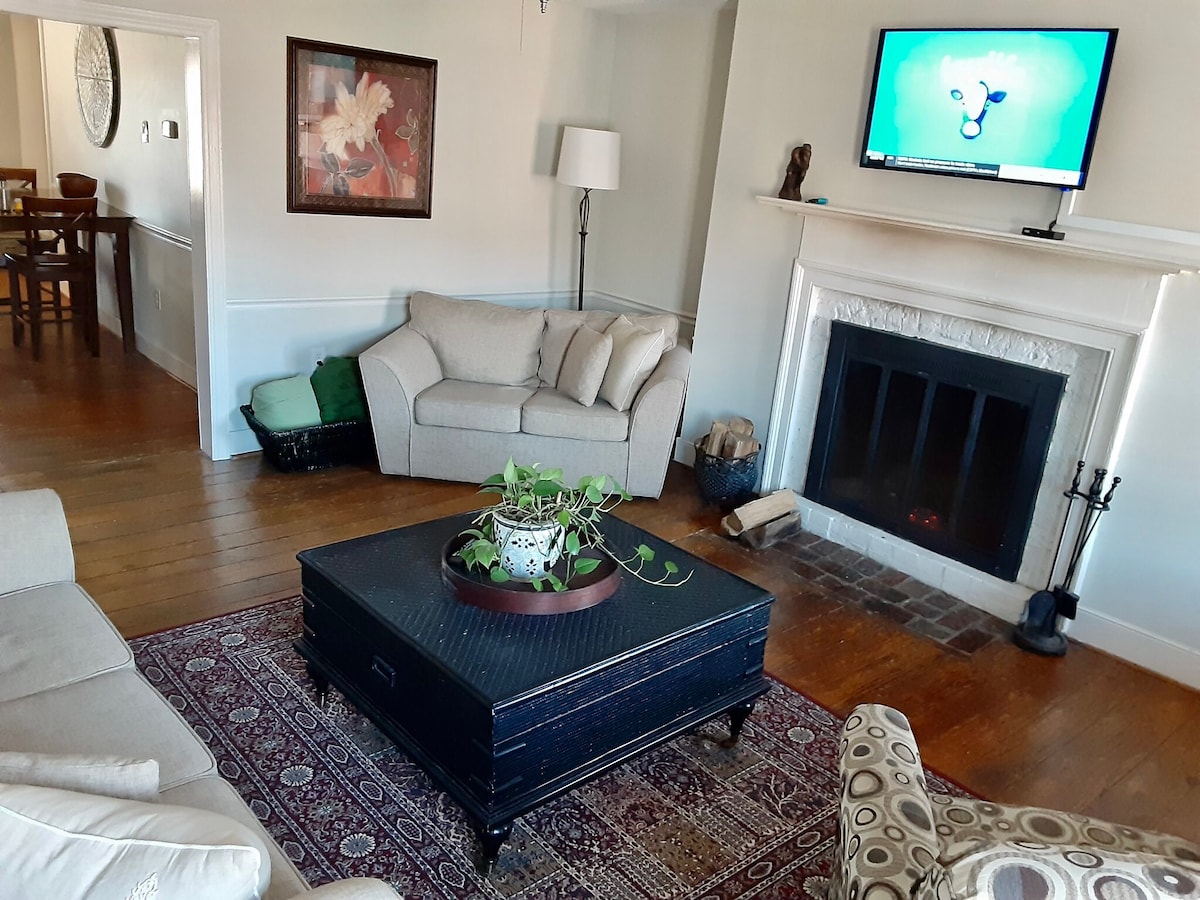 A living area features a comfortable beige sofa and an accent chair, arranged around a dark coffee table with a plant centerpiece. A TV is mounted on the wall above a traditional fireplace. Natural light streams through a nearby window, illuminating the hardwood floors.