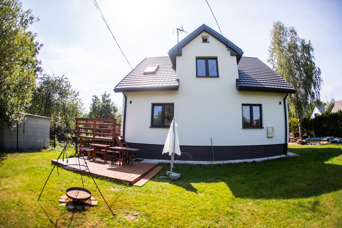 The exterior of a two-story home is viewed from the yard, featuring a neatly maintained lawn. A wooden deck with seating is present, complemented by an outdoor umbrella and a fire pit. The house showcases large windows and a simple, modern design.
