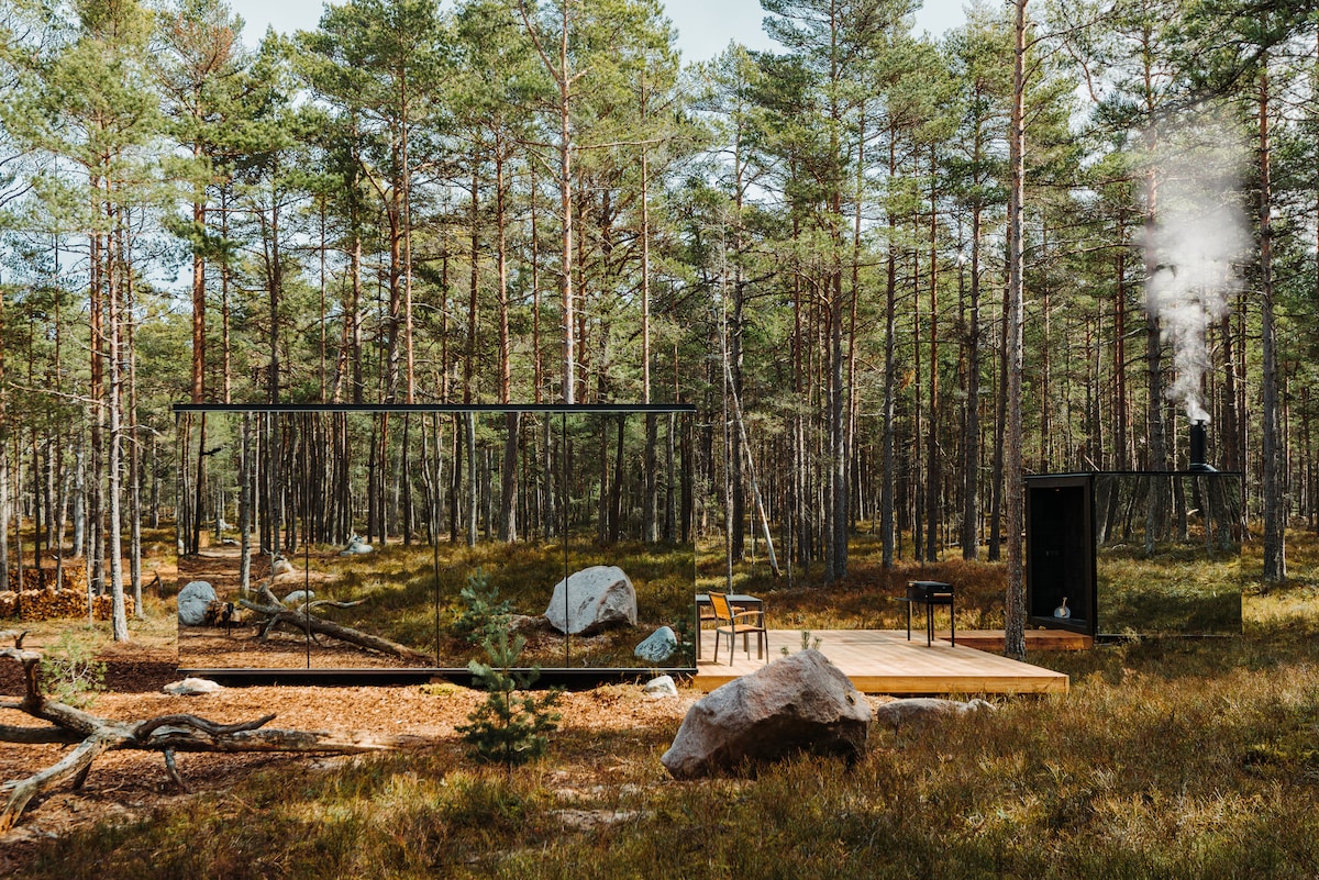 A modern cabin is nestled in a forest setting, surrounded by tall pine trees. The design incorporates large glass panels, reflecting the natural surroundings. A wooden deck is visible, along with a small outdoor area featuring rustic seating. Smoke gently rises from a nearby outdoor sauna.
