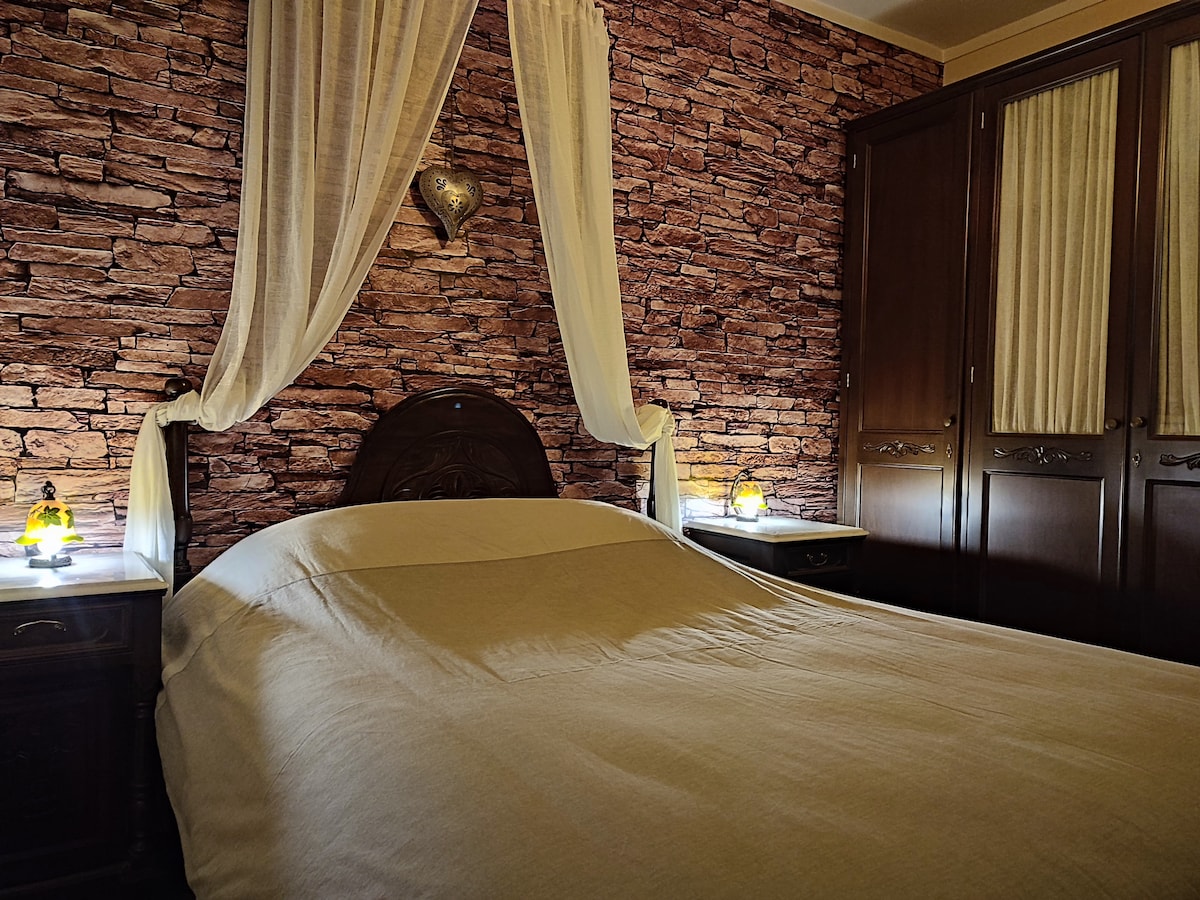 A spacious bed is positioned against a stone-patterned wall, complemented by soft, draping curtains. Nightstands with lamps on either side provide gentle lighting, while a wooden wardrobe stands nearby, featuring detailed woodwork and glass-paneled doors.