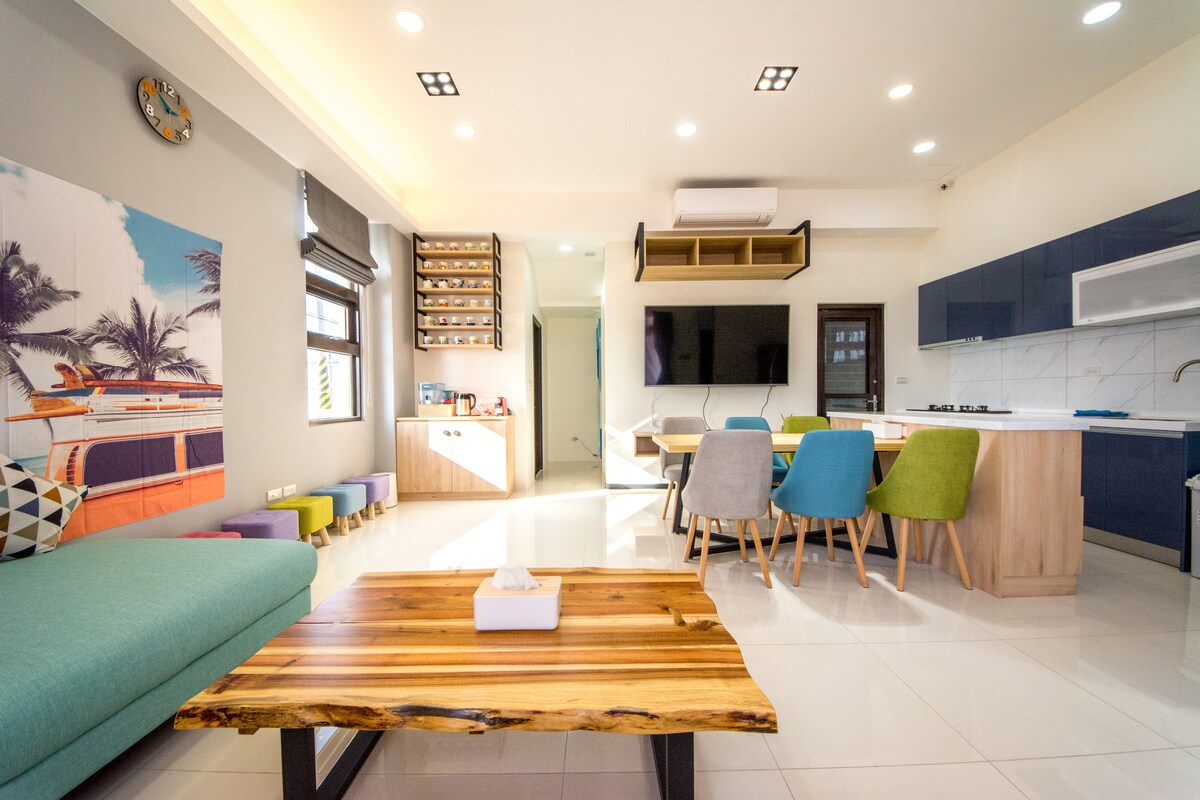 An open-plan living space features a vibrant blue sofa and a wooden coffee table at the center. A modern kitchen with sleek cabinetry is visible, along with a dining area that includes a round table surrounded by colorful chairs. Bright light floods in from the windows.