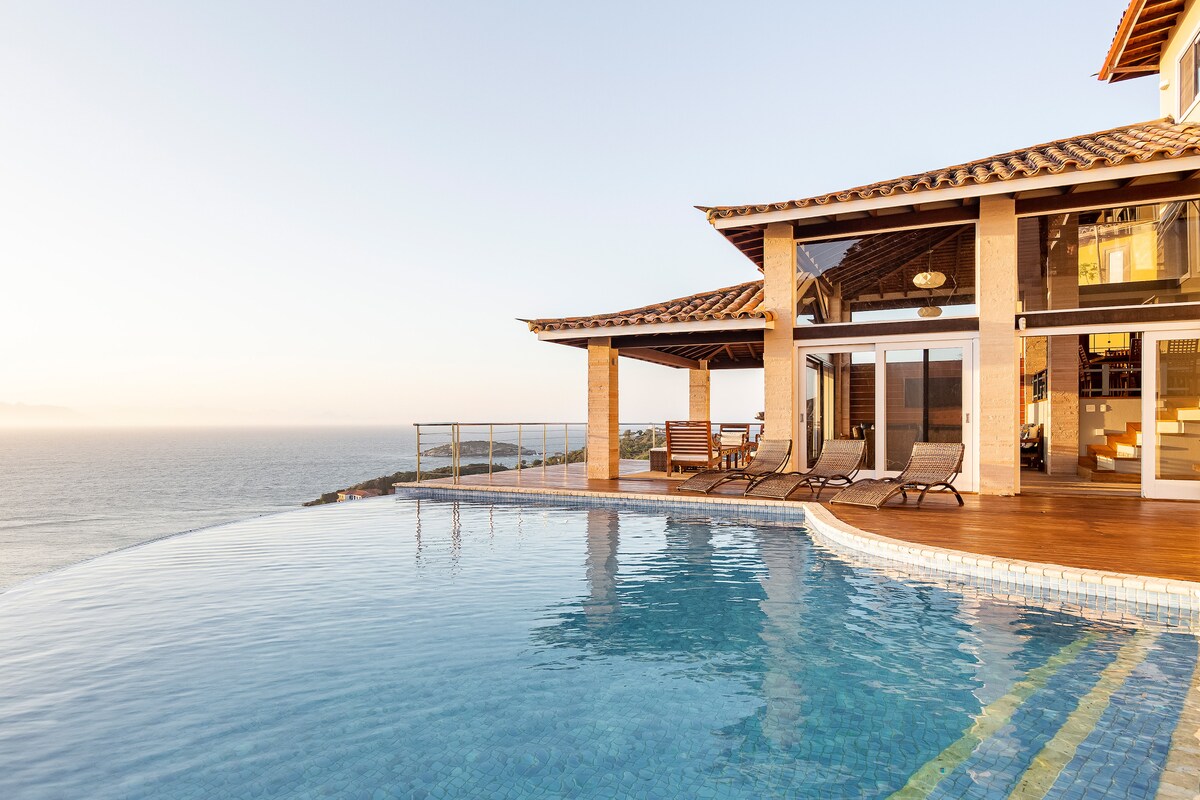 An infinity pool glistens in the sunlight, blending seamlessly with the horizon. The elegant house features large windows and a spacious terrace, offering panoramic views of the ocean. Comfortable lounge chairs are positioned nearby, inviting relaxation by the water.