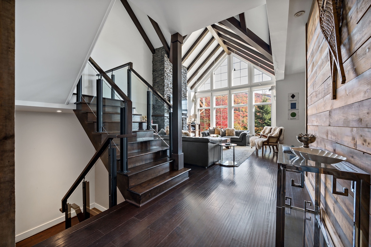 An open staircase leads to an upper level, framed by a striking feature wall of wood and stone. Large windows allow natural light to fill the spacious living area, showcasing comfortable seating and seasonal views of vibrant foliage.
