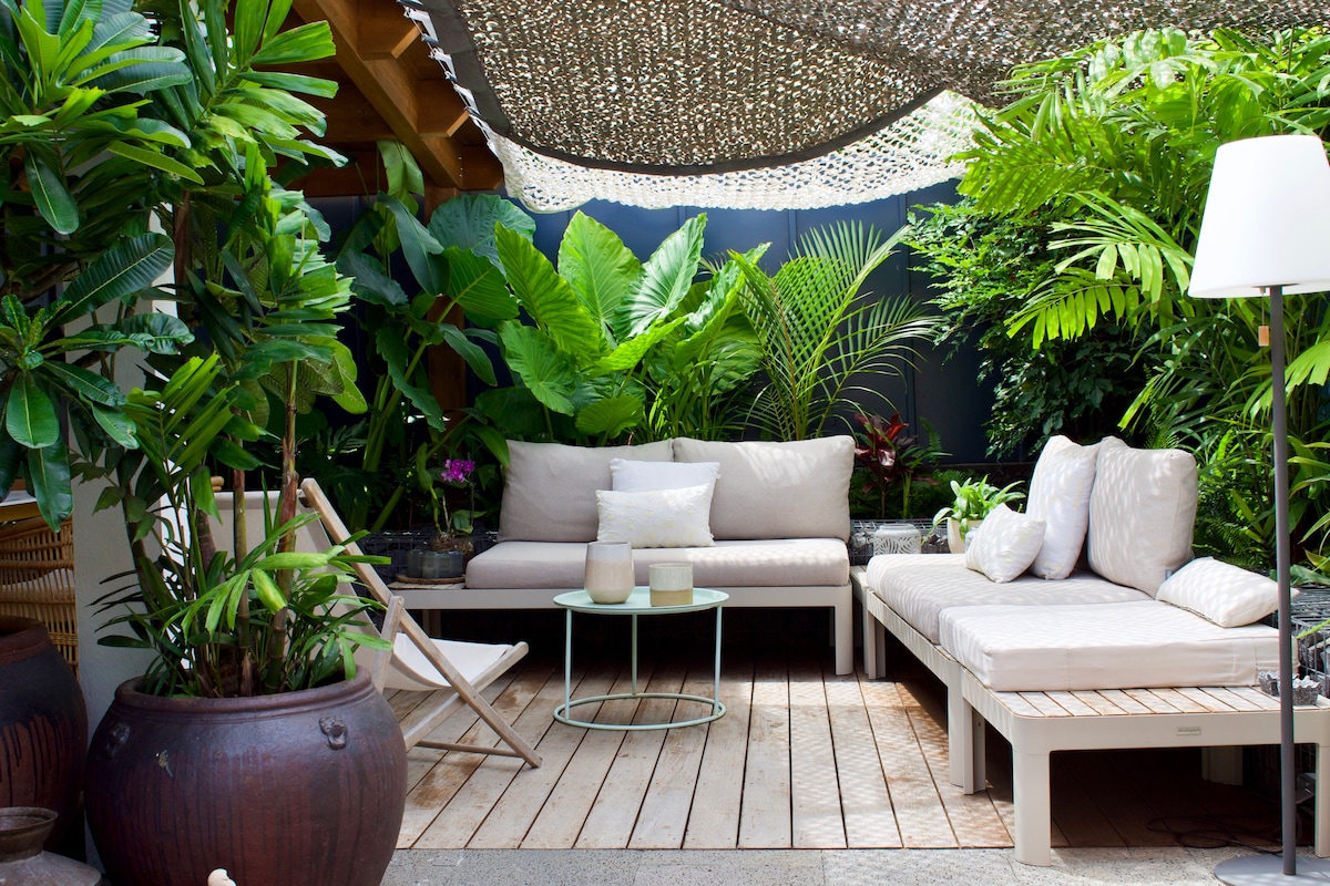 A serene outdoor seating area is surrounded by lush green plants, creating a private retreat. Comfortable sectional sofas paired with a glass coffee table are placed on wooden decking. A folding chair offers additional seating under a textured canopy, providing shaded comfort.
