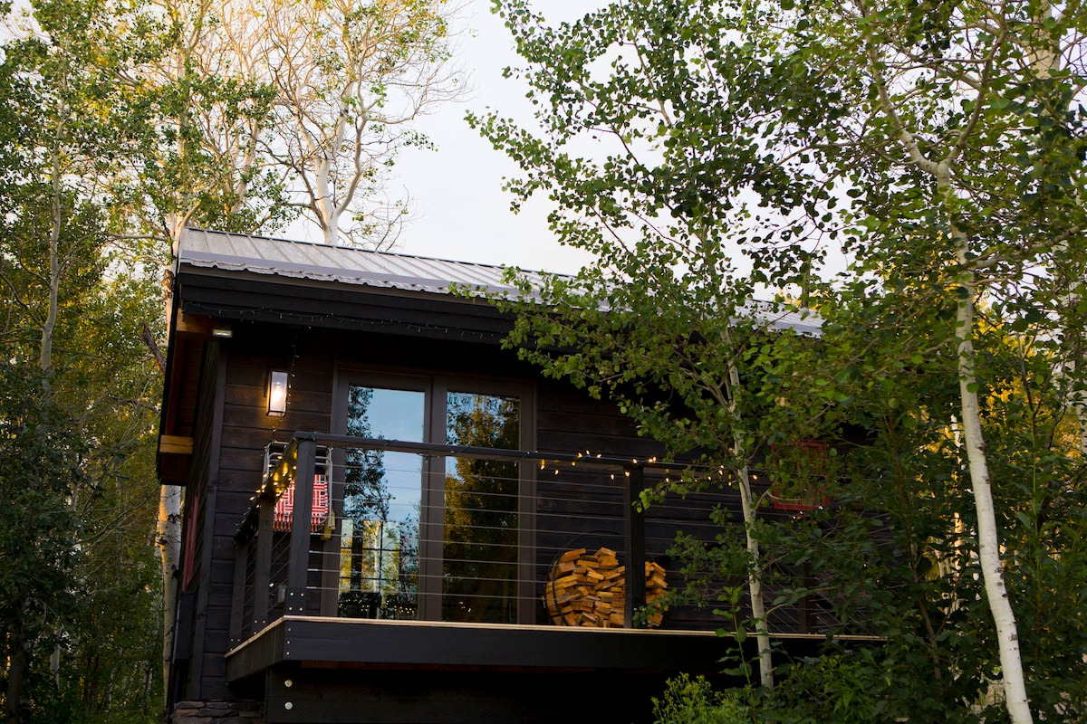 The handcrafted timber frame cabin is surrounded by lush greenery, showcasing large windows that invite natural light. A spacious deck is visible, adorned with decorative lights, while neatly stacked firewood is positioned nearby, enhancing the rustic charm of the retreat.