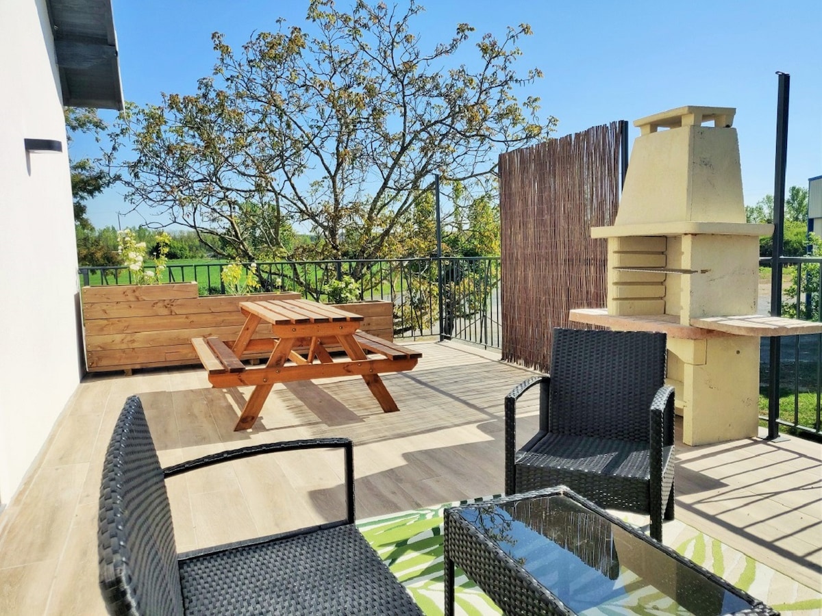 A spacious terrace features a wooden picnic table and two wicker chairs arranged around a glass-topped table. A built-in barbecue stands nearby, surrounded by greenery and a clear blue sky. The area offers a comfortable space for outdoor relaxation and dining.