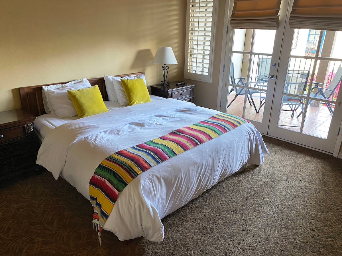 A spacious sleeping area features a large bed adorned with a colorful blanket across the foot. Two bright yellow pillows are positioned against the white bedding. Natural light filters through the balcony doors, which open to a cozy outdoor seating area.
