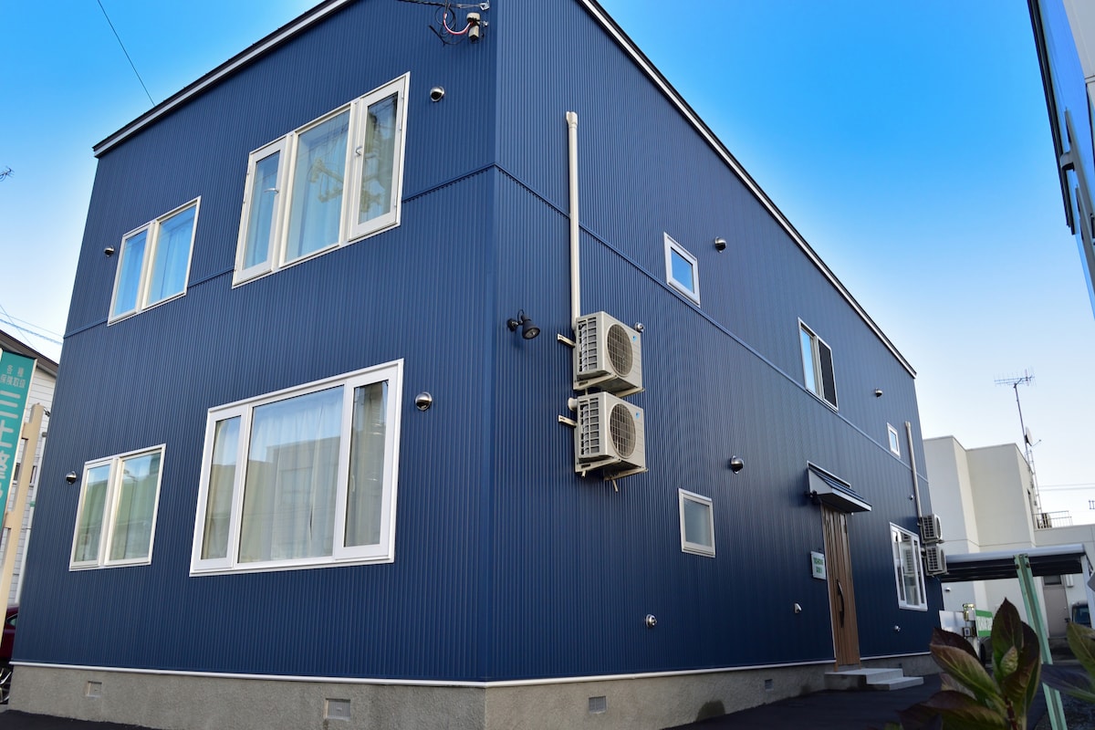 The exterior of a modern blue apartment building is featured, showcasing multiple windows of various sizes. Air conditioning units are mounted on the side, and a wooden door with a small awning adds a welcoming touch. The surrounding area is paved.