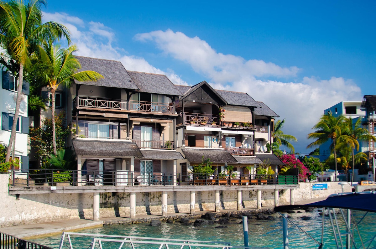 A beachfront penthouse is seen with multiple levels, featuring a mix of dark wooden and light-colored exteriors. Palm trees line the property, and vibrant flowers add color. A small dock is visible in the foreground, with clear water gently lapping against the structure.