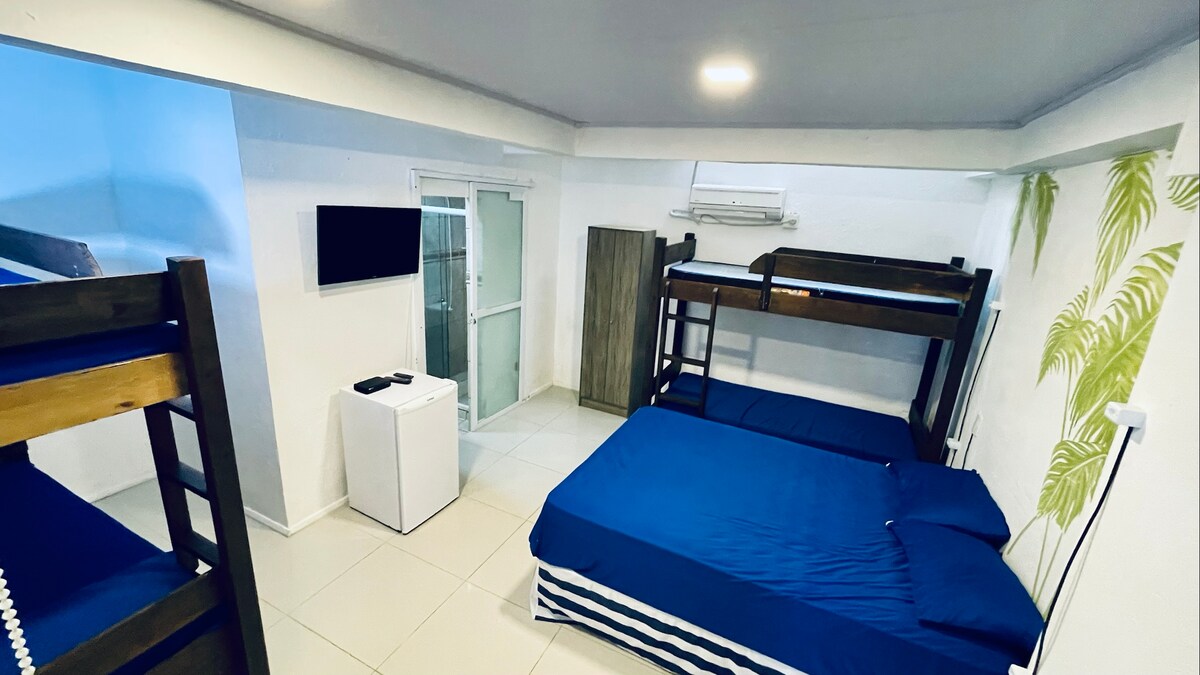 A well-arranged sleeping area features a spacious bed and two bunk beds, all made with blue bedding. A small refrigerator is positioned nearby, alongside a wall-mounted TV. Air conditioning is installed, and natural light enters through a window adorned with a light curtain.