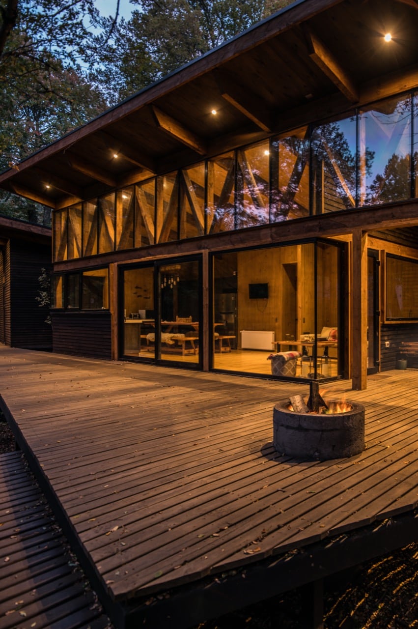 A modern cabin is presented with large glass panels, reflecting the surrounding forest. A wooden deck extends outward, featuring an outdoor fire pit. The warm light from inside enhances the inviting ambiance of the communal space visible through the glass.