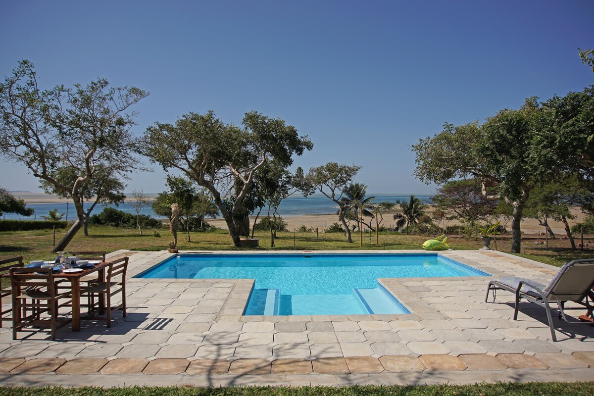 A clear blue swimming pool is surrounded by stone paving and overlooks a scenic view of the beach and ocean. A dining table and chairs are set nearby, shaded by trees, creating a relaxing outdoor space.