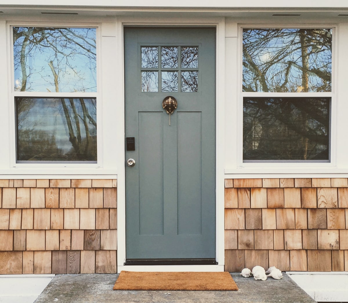 The entrance features a blue front door with a circular door knocker, framed by two large windows on each side. The exterior is adorned with warm wooden shingles, complemented by a welcome mat at the foot of the door.