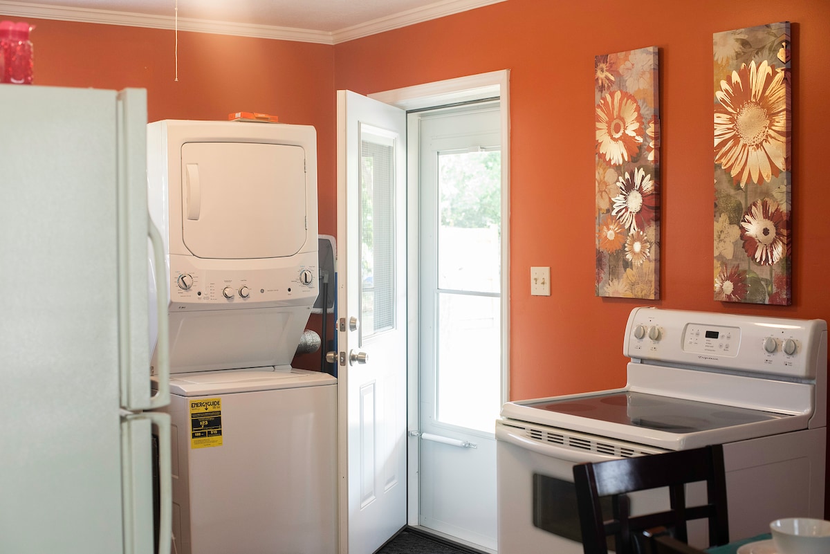 A laundry area features a stacked washer and dryer alongside a door leading to the outside. Two floral wall art pieces add a touch of color to the warm orange walls. A modern range is positioned next to the washer and dryer, creating a functional layout.