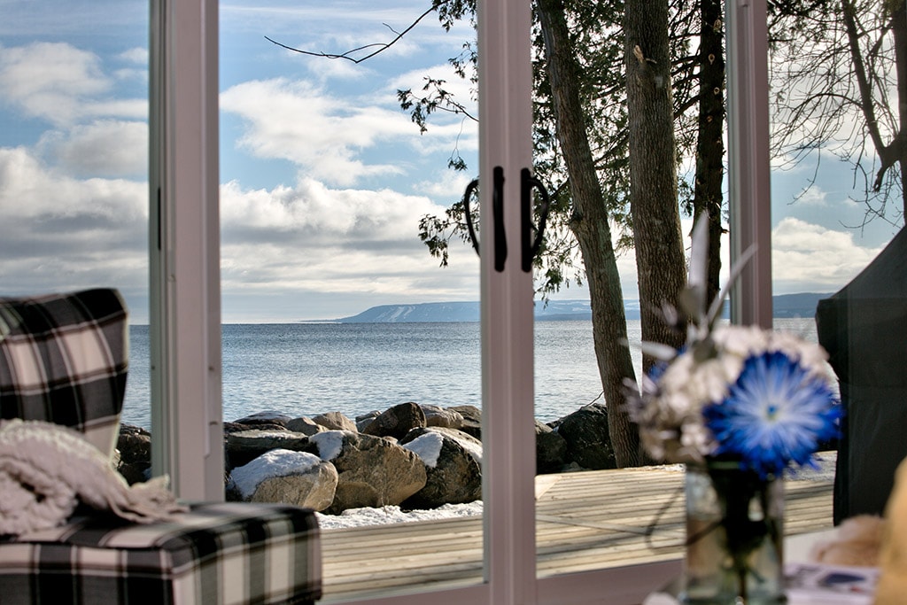A view from inside the cottage is framed by large glass doors, showcasing the serene waters of Georgian Bay against a backdrop of distant hills. Natural light floods the interior, illuminating a cozy seating arrangement with checkered upholstery and decorative flowers on a nearby table.