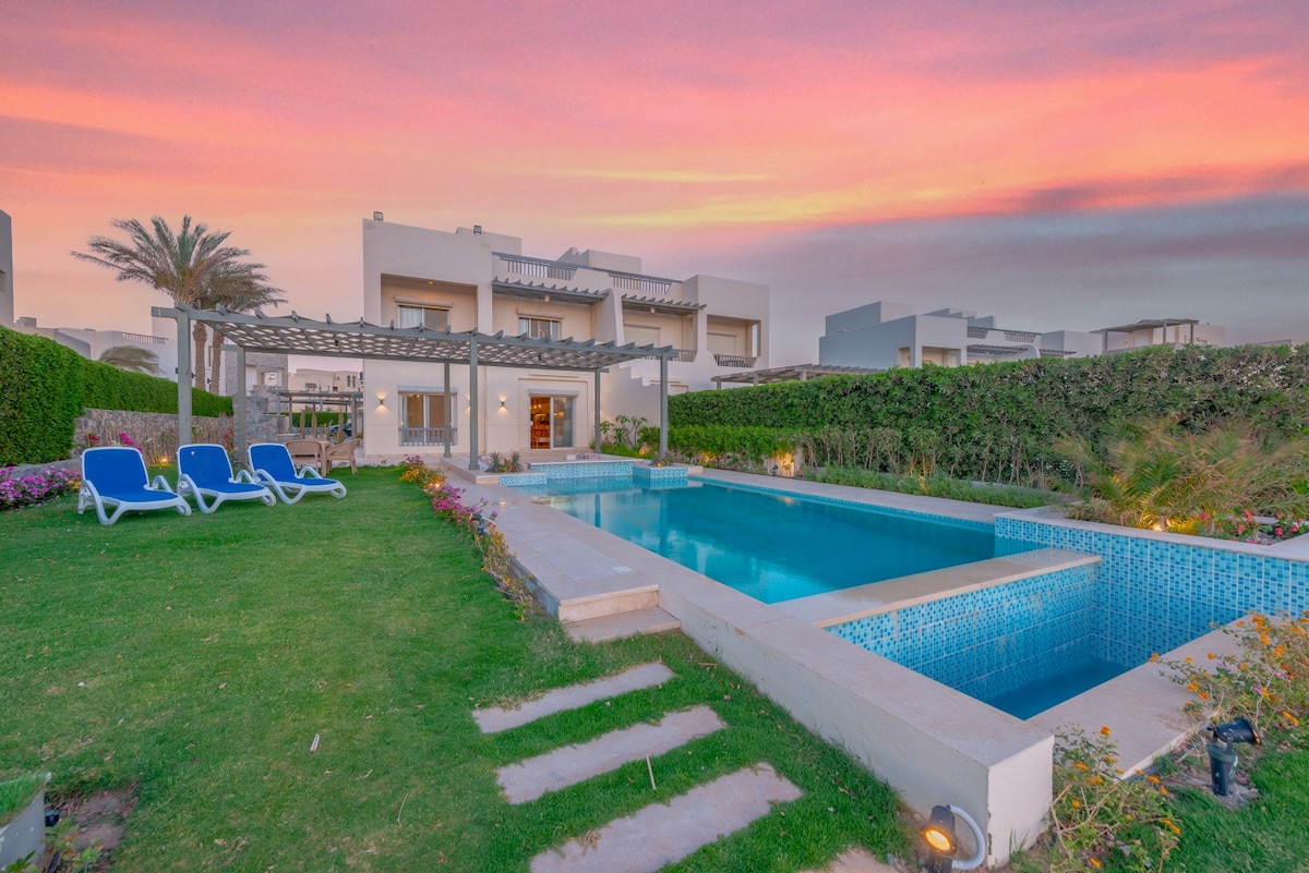 The image captures a modern villa surrounded by lush greenery, featuring a private swimming pool and an inviting garden area. Comfortable lounge chairs are placed next to the pool, with a pergola offering shade. The sky displays vibrant colors at dusk, enhancing the serene atmosphere.