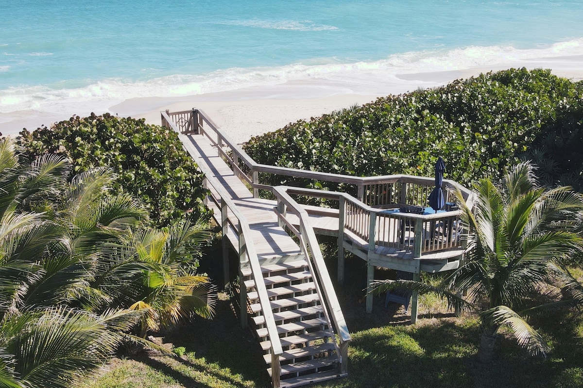A wooden crossover path leads through lush greenery to the beach. Seating is provided at the top of the steps, offering a view of the turquoise ocean and sandy shore. The scene is framed by tropical foliage, enhancing the connection to nature.