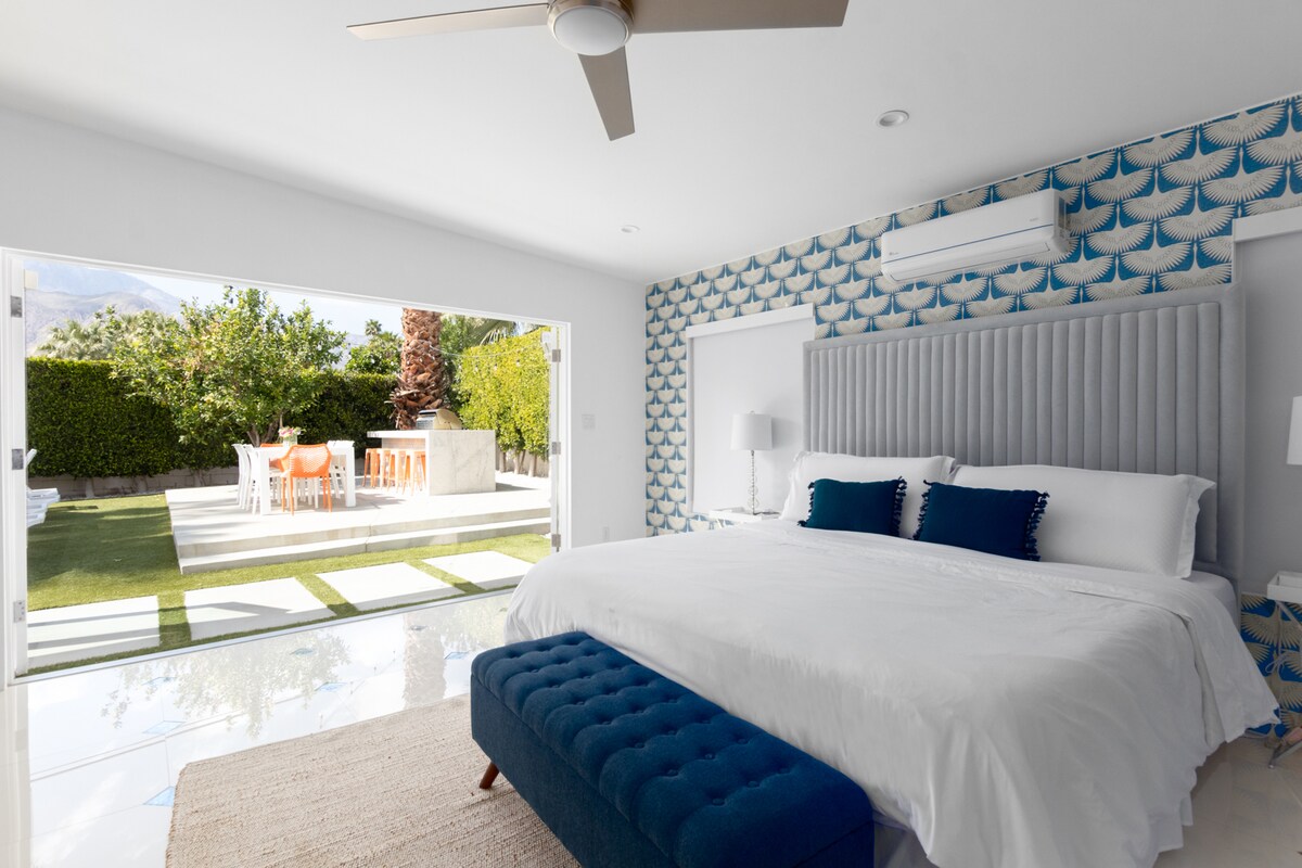 A spacious bedroom features a king bed with luxury bedding and a tufted blue bench at its foot. Natural light floods in through double doors that open to a beautifully landscaped backyard with outdoor dining. Decorative wallpaper adds a modern touch to the room.