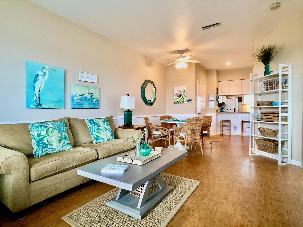 A spacious living area features a comfortable sofa adorned with tropical-themed pillows, complemented by a stylish coffee table. Light-colored walls are decorated with ocean-inspired artwork. A dining area with wicker chairs is visible, leading to a well-equipped kitchen with white cabinetry.