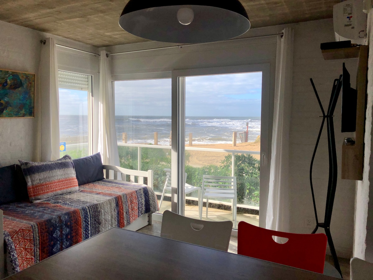 A living area is shown, featuring a sofa with colorful textiles, adjacent to large glass windows offering views of the beach and ocean. Natural light fills the space, complemented by white curtains. A dining table is positioned nearby, enhancing the open atmosphere.