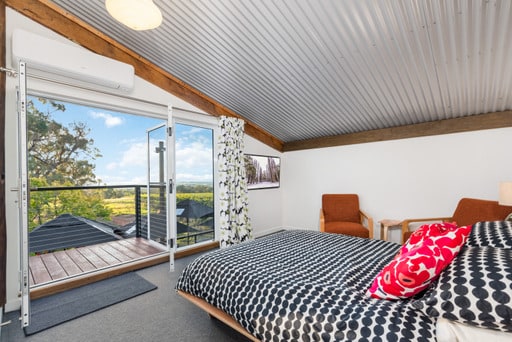 A modern loft space features a comfortable bed with patterned bedding. Two armchairs are positioned nearby, and large doors open to a balcony overlooking the vineyard. Natural light fills the room, complemented by wood accents and a contemporary feel.