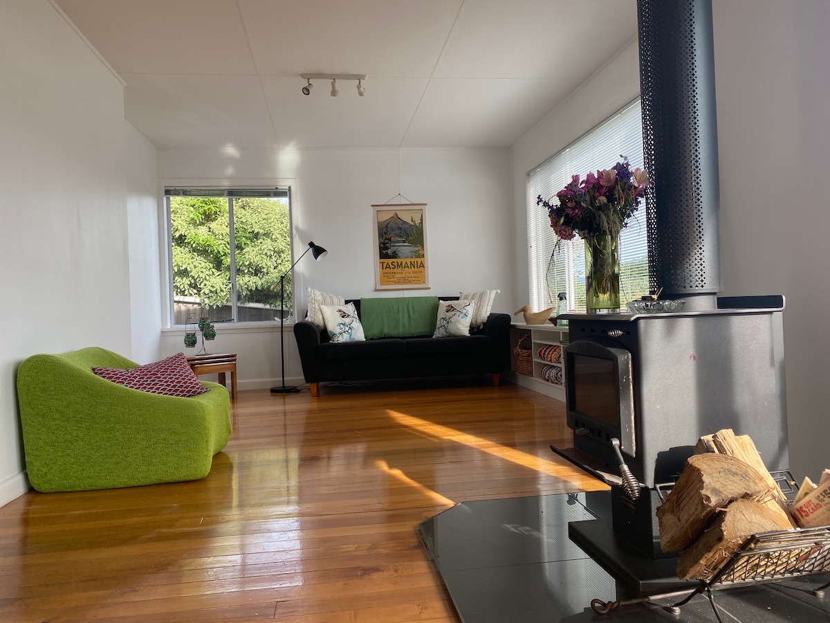 A bright living area features large windows allowing natural light to fill the space. A black couch is complemented by a vibrant green chair and a vase of flowers. A wood stove is positioned beside a stack of firewood, enhancing the cozy setting.