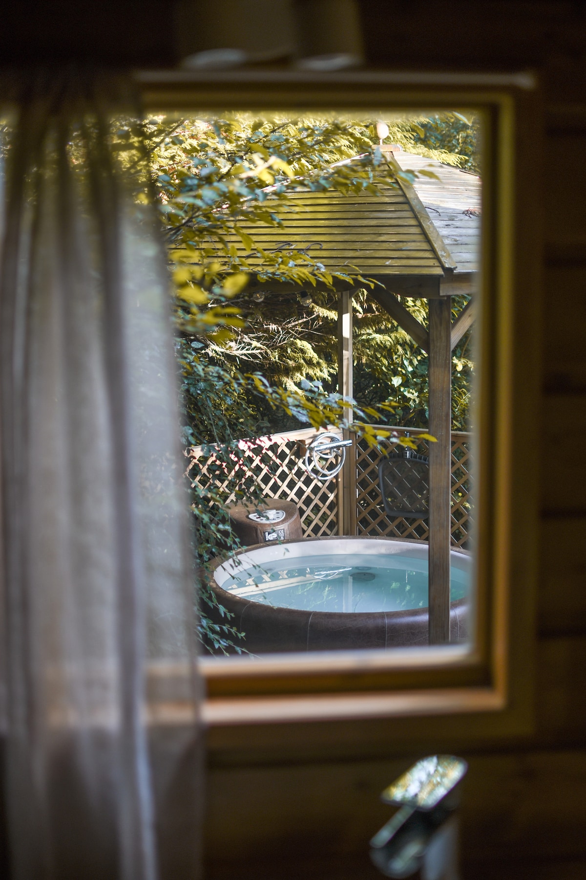 A view from a window showcases a private hot tub, nestled under a wooden shelter, surrounded by lush greenery. Sunlight filters through the leaves, creating a tranquil and serene atmosphere. The inviting waters of the hot tub reflect the natural surroundings.