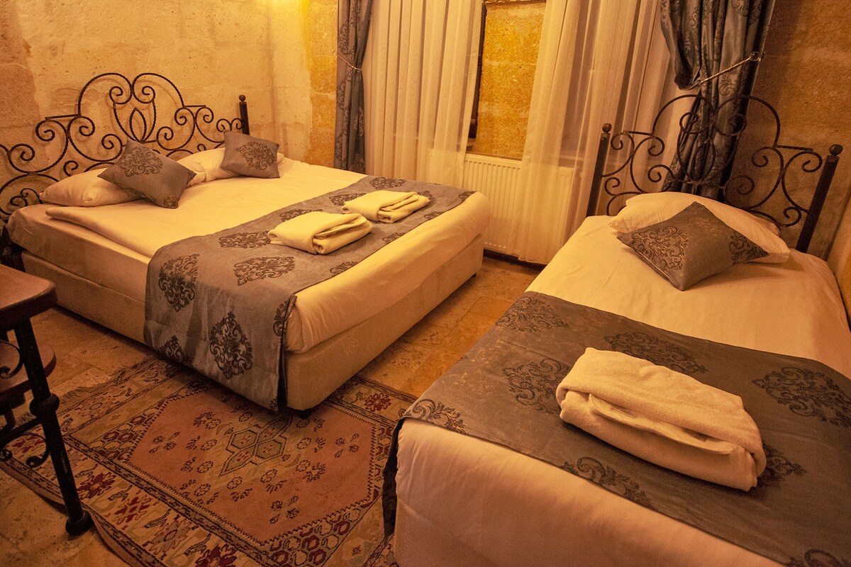 A warm sleeping area features two spacious beds with decorative blankets and pillows. Soft lighting creates a cozy ambiance. A wooden side table is positioned beside one bed, and a patterned area rug adds comfort to the stone floor.