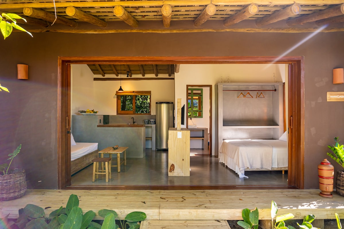 An open concept space is presented, featuring a kitchen area with modern appliances, a wooden dining table, and two sleeping areas. The queen size bed and a single bed are visible, with natural light flooding the interior through large glass doors that connect to the outdoor area.