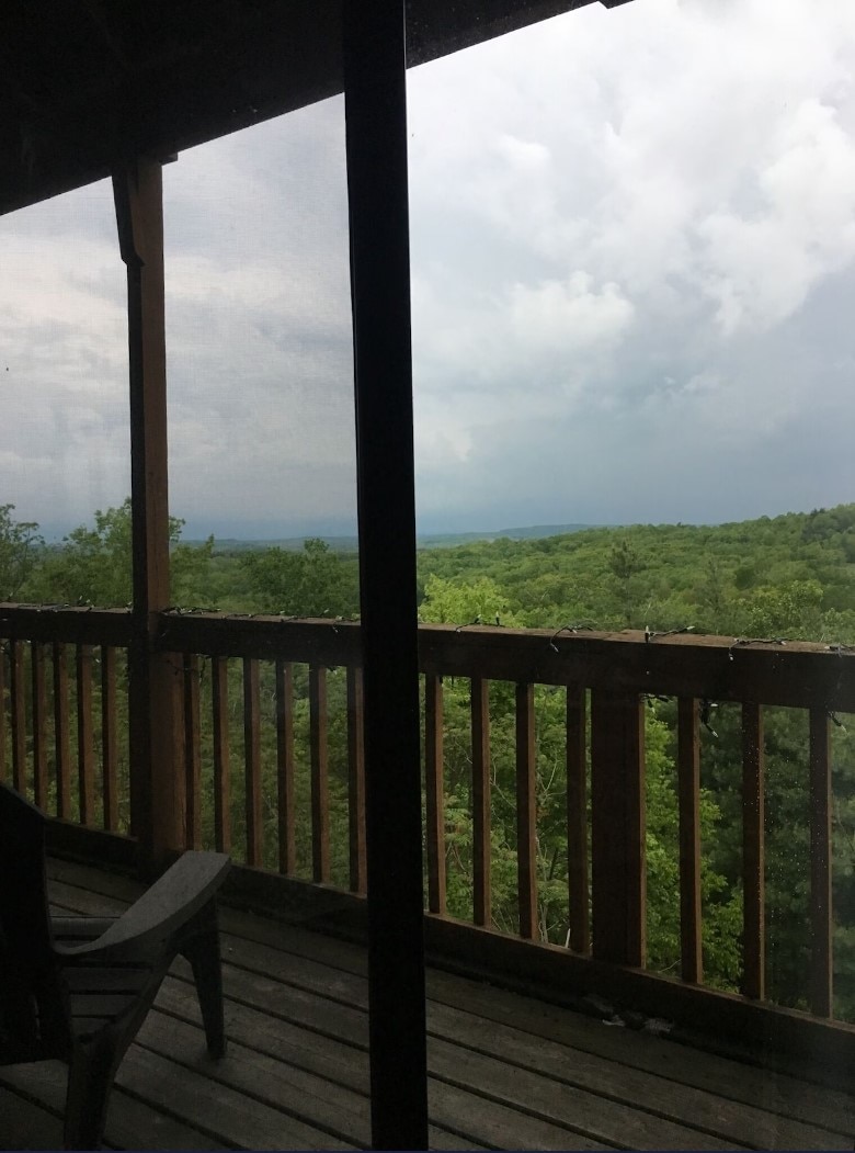 A screened balcony view reveals lush greenery stretching into the distance beneath a cloudy sky. The wooden railing adds a rustic touch to the serene outdoor space, inviting guests to enjoy the natural surroundings.