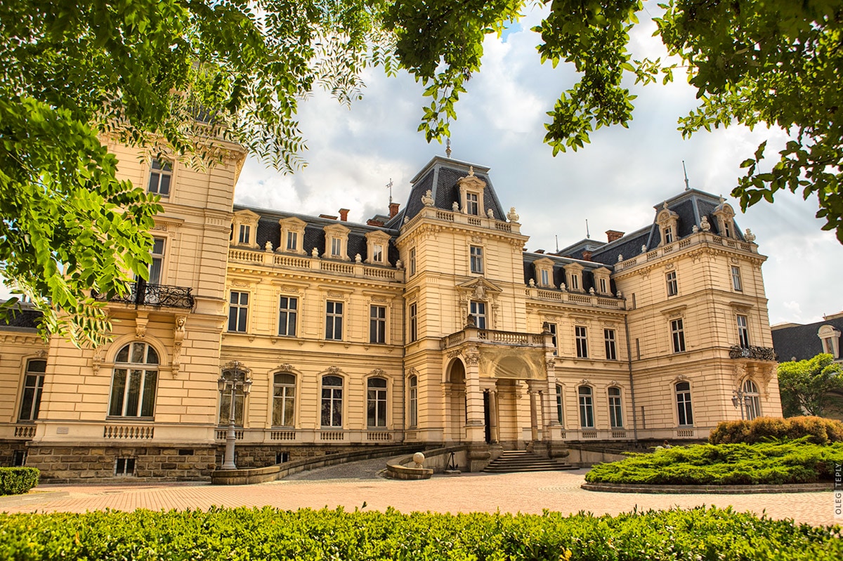 A grand mansion with intricate architectural details is surrounded by lush greenery. The facade features multiple stories, large windows, and decorative balconies, showcasing elegant design. A neatly paved area in front offers an unobstructed view of the building's classic charm.