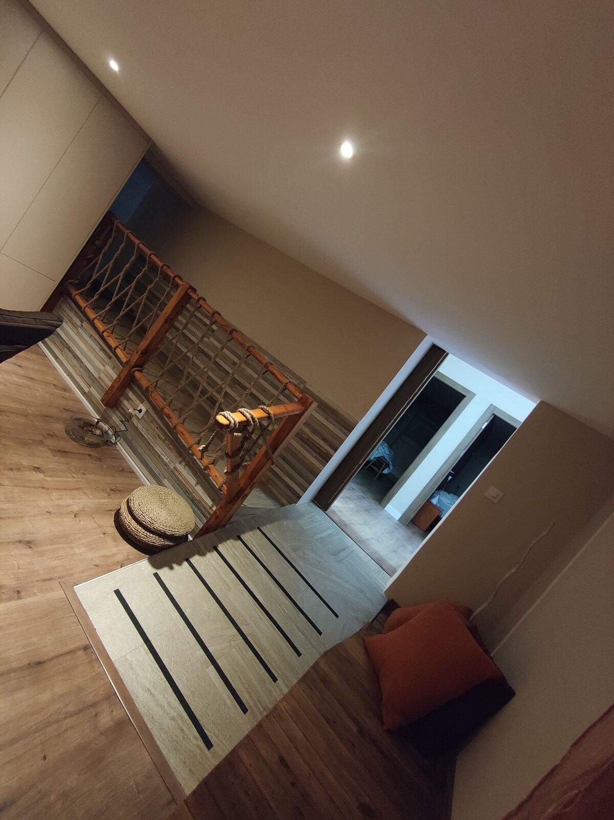 A spacious hallway is viewed, featuring wooden flooring and soft lighting. A woven basket is positioned on the floor, and a cozy rug with a striped pattern lies near an inviting seating area with cushions. A railing adds a subtle touch of warmth to the space.