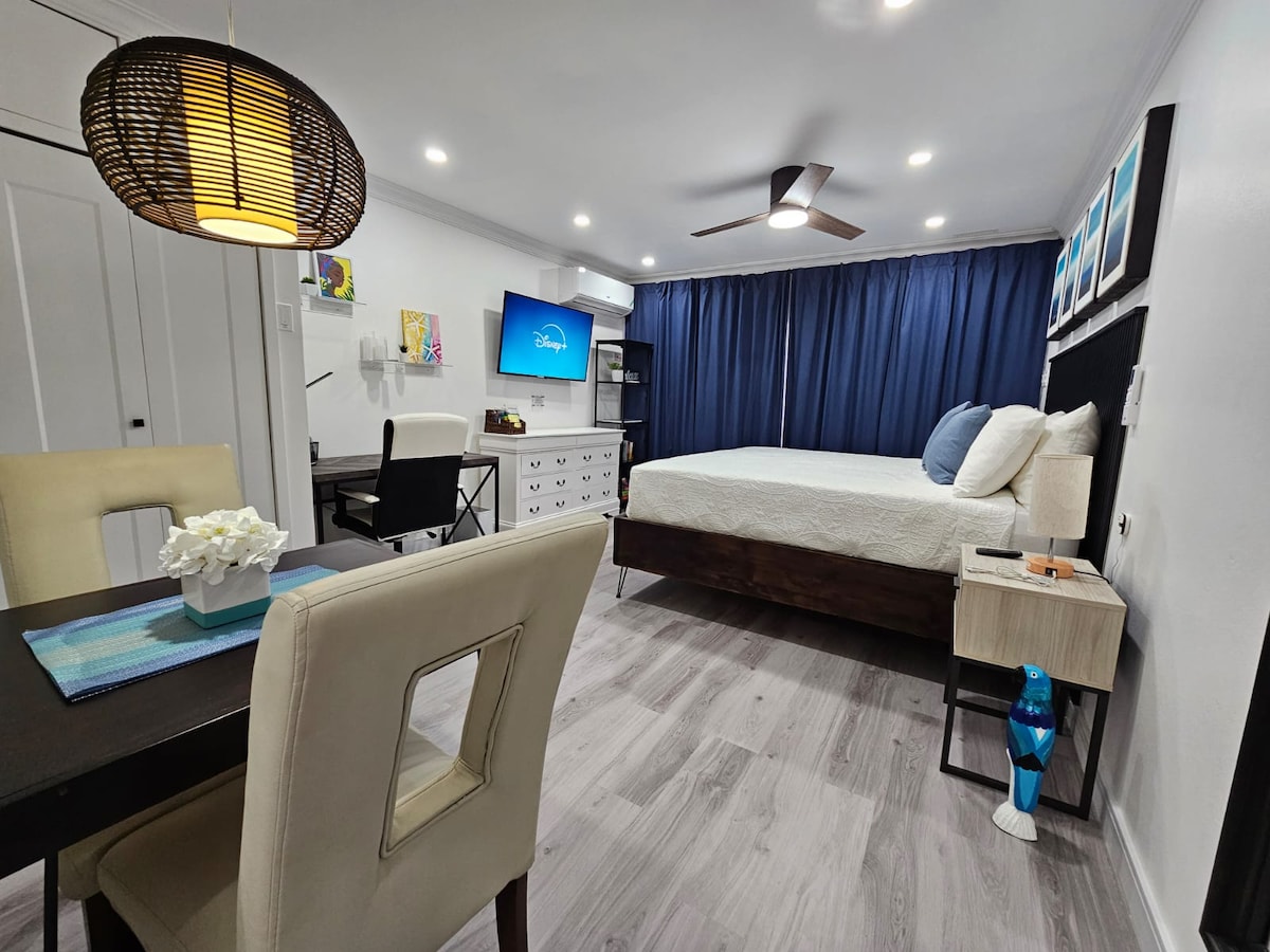 The studio condo features a modern design with a spacious bed facing a wall-mounted smart TV. A dining table for two and a comfortable work desk are positioned nearby, illuminated by a stylish pendant light. Navy blue curtains add a touch of elegance to the room.