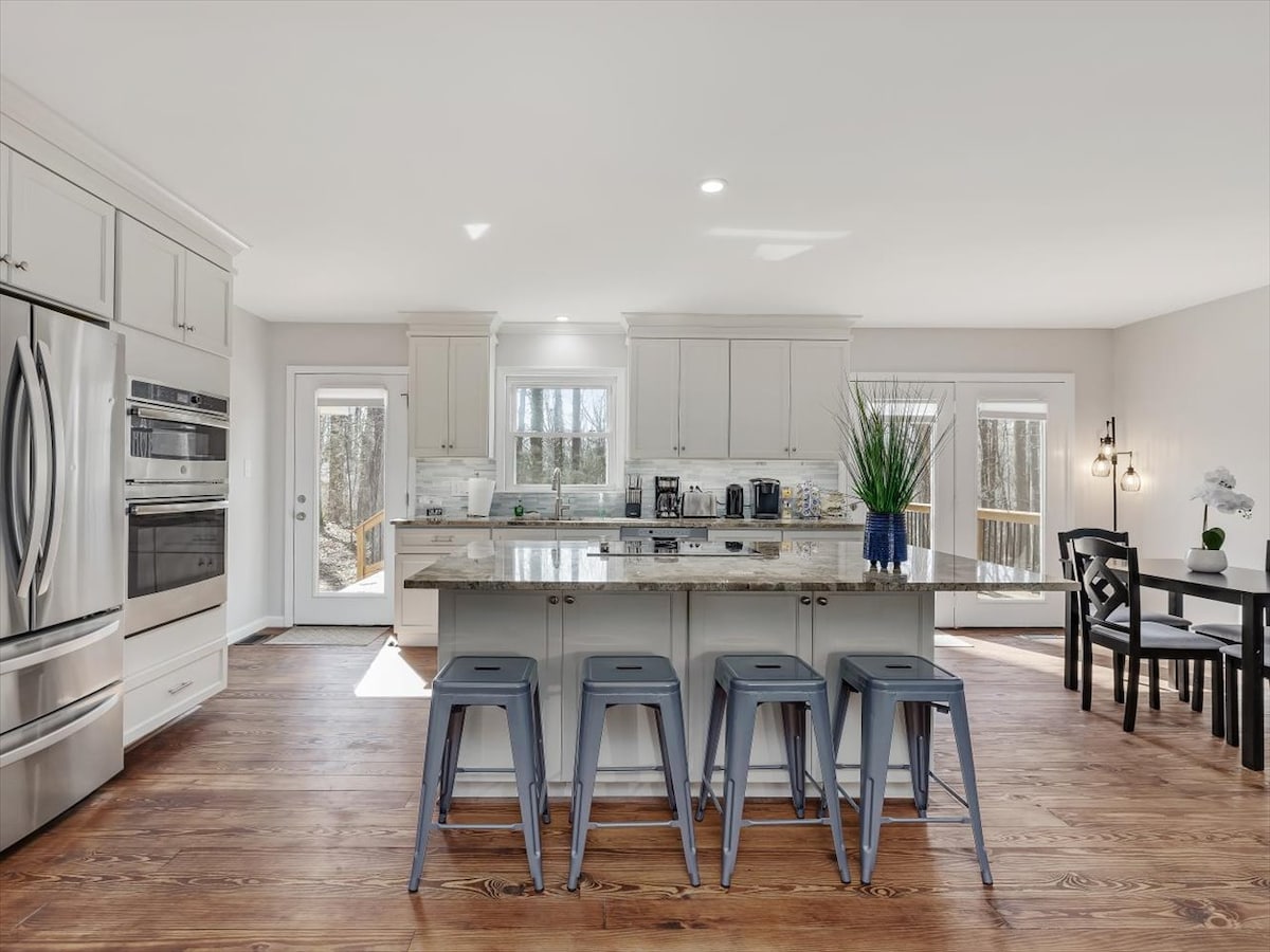A modern kitchen features a spacious island with metallic stools and high-end appliances. White cabinetry contrasts with the warm hardwood flooring. Natural light floods the space through multiple windows, highlighting the stylish countertops and an adjoining dining area with a dark table and seating.
