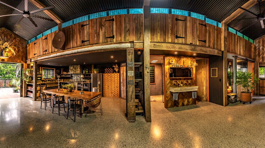 An open-plan living space is characterized by rustic wooden beams and a polished concrete floor. A large dining table occupies the center, surrounded by chairs. A well-equipped kitchen is visible to the side, with natural light flooding the area from large windows and doors.