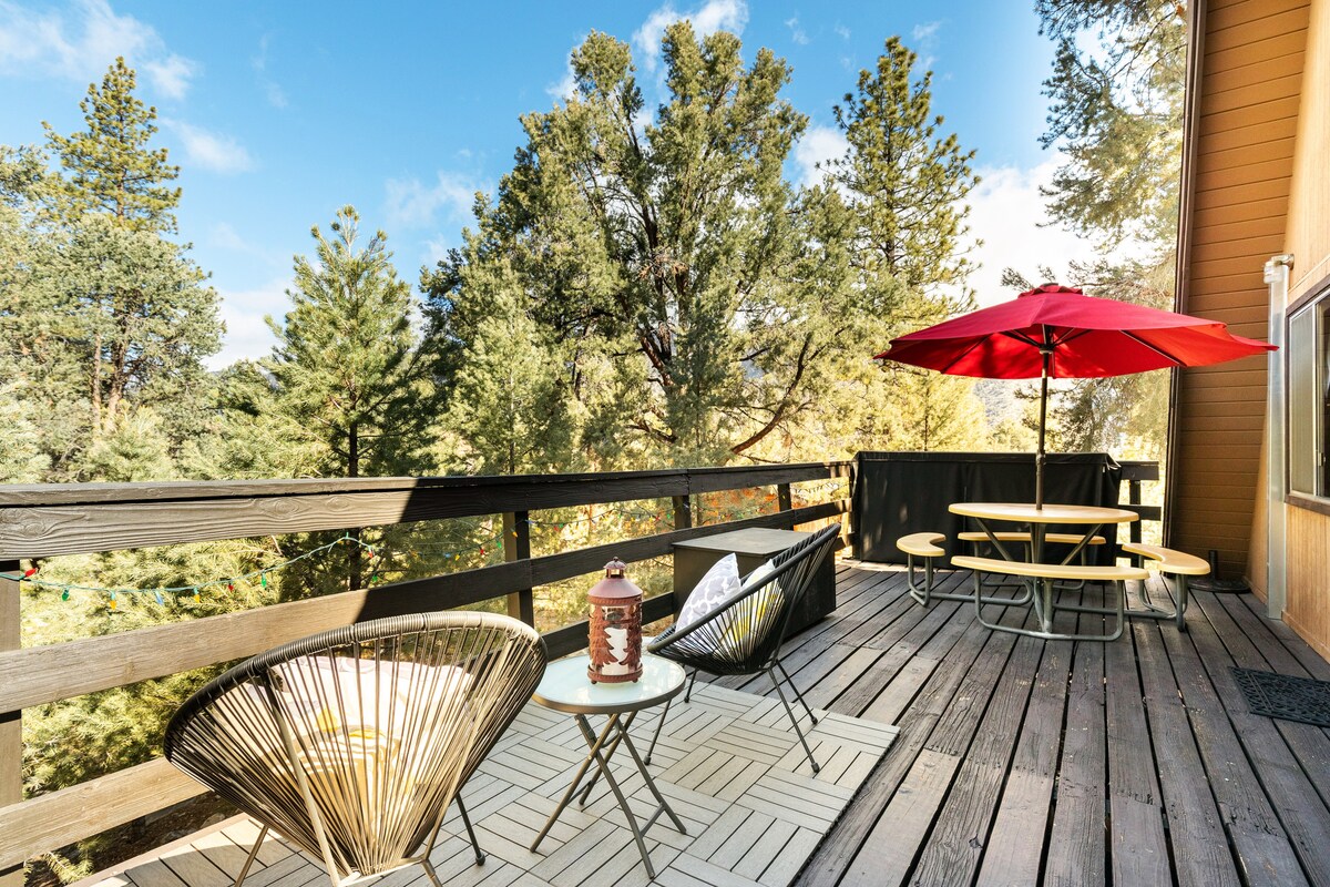 A spacious deck features a picnic table surrounded by trees, providing a serene outdoor setting. Two black, woven chairs are positioned beside a small table, while a red patio umbrella offers shade. The wood decking adds a rustic touch, complementing the natural surroundings.